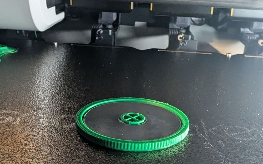 A half-printed green coin on the 3D printer bed with a steel washer set inside it, print heads above